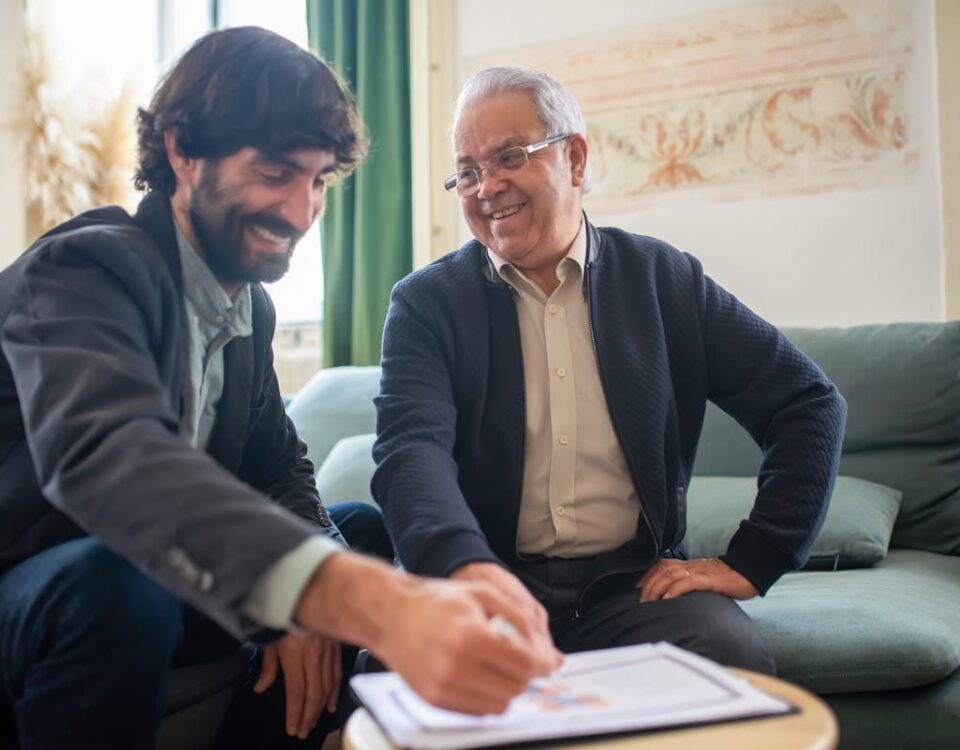 Como a tecnologia facilita a compra de imóveis hoje? Two Men Smiling While Discussing Paperwork on a Wooden Table