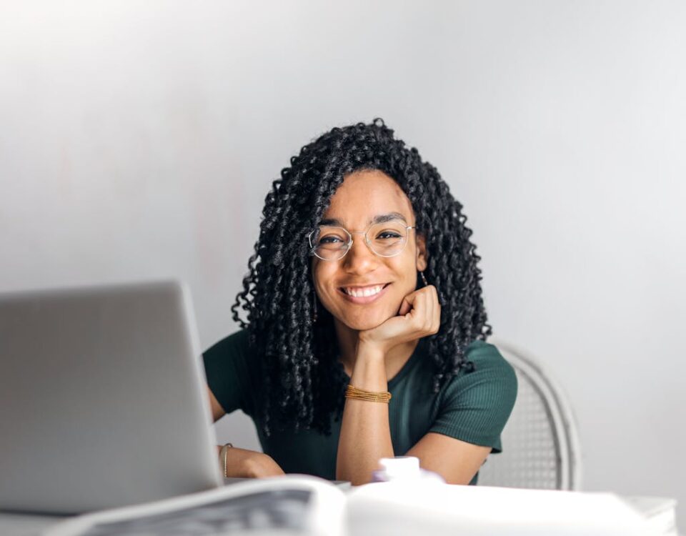 Apps que revolucionam seu método de estudo Joyful businesswoman with curly hair smiling at camera while using laptop indoors.