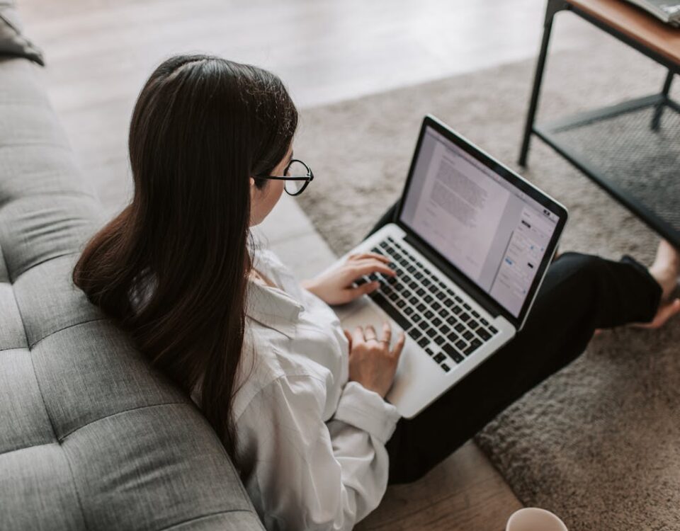 Como escolher um curso online em tecnologia? Woman working remotely with a laptop on the floor next to a sofa, enjoying comfortable home office setup.