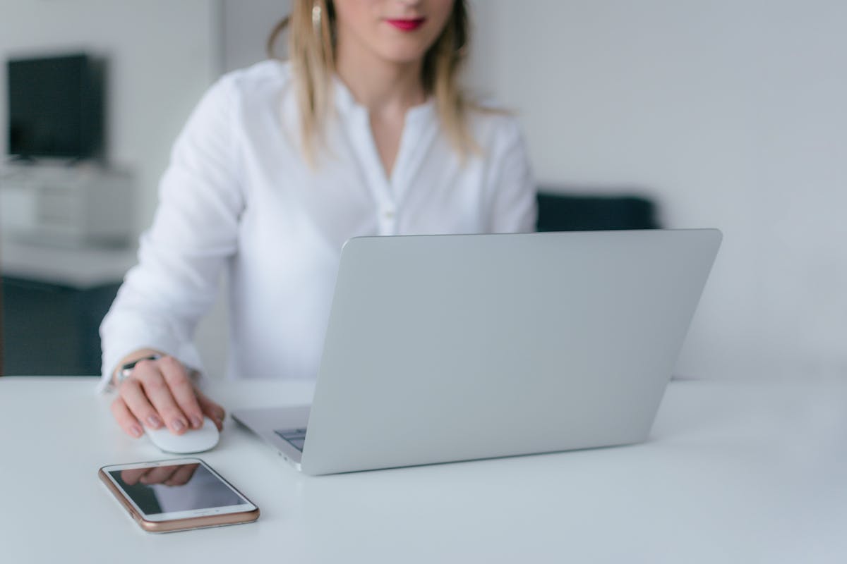 Ferramentas digitais para auxiliar administrativos A woman working at a desk using a laptop and smartphone, exemplifying remote work.
