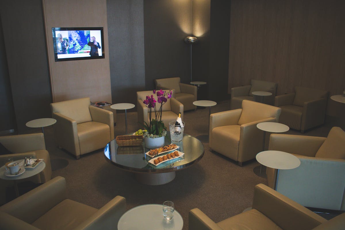 Como funcionam os cartões que oferecem acesso às salas VIP? Elegant airport lounge with beige leather seats and orchid decor, offering comfort and luxury food service.