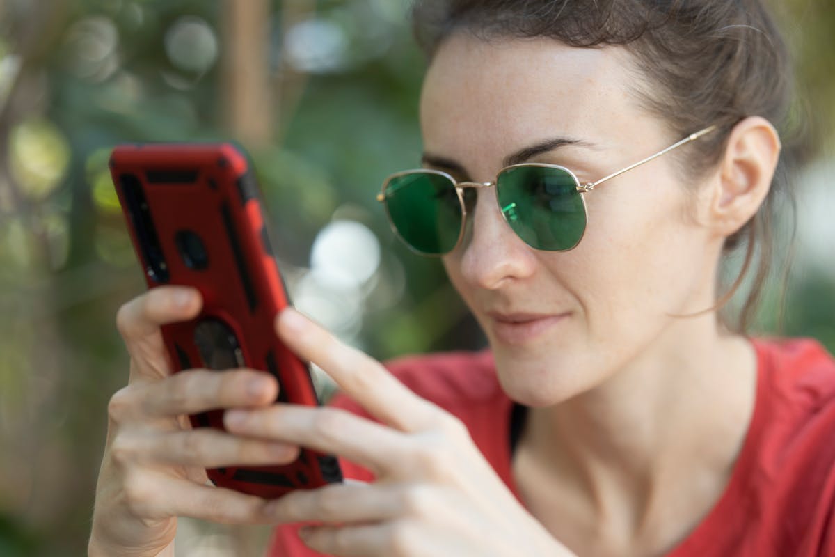 Adult woman using her smartphone while wearing sunglasses, enjoying a sunny day outdoors.