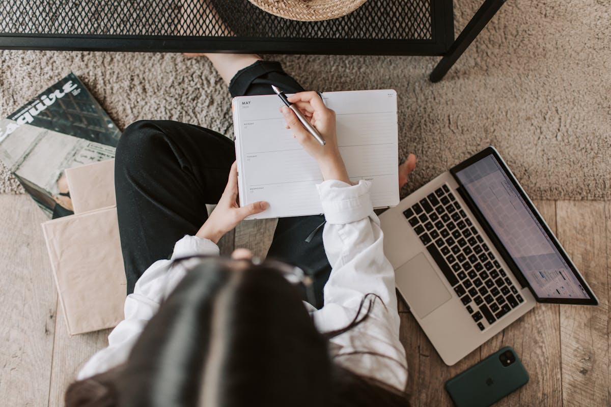 Aplicativos que ajudam no estudo para provas da CNH Top view of anonymous woman in casual wear sitting on floor with laptop and smartphone and creating plan on notebook while resting during break in modern living room