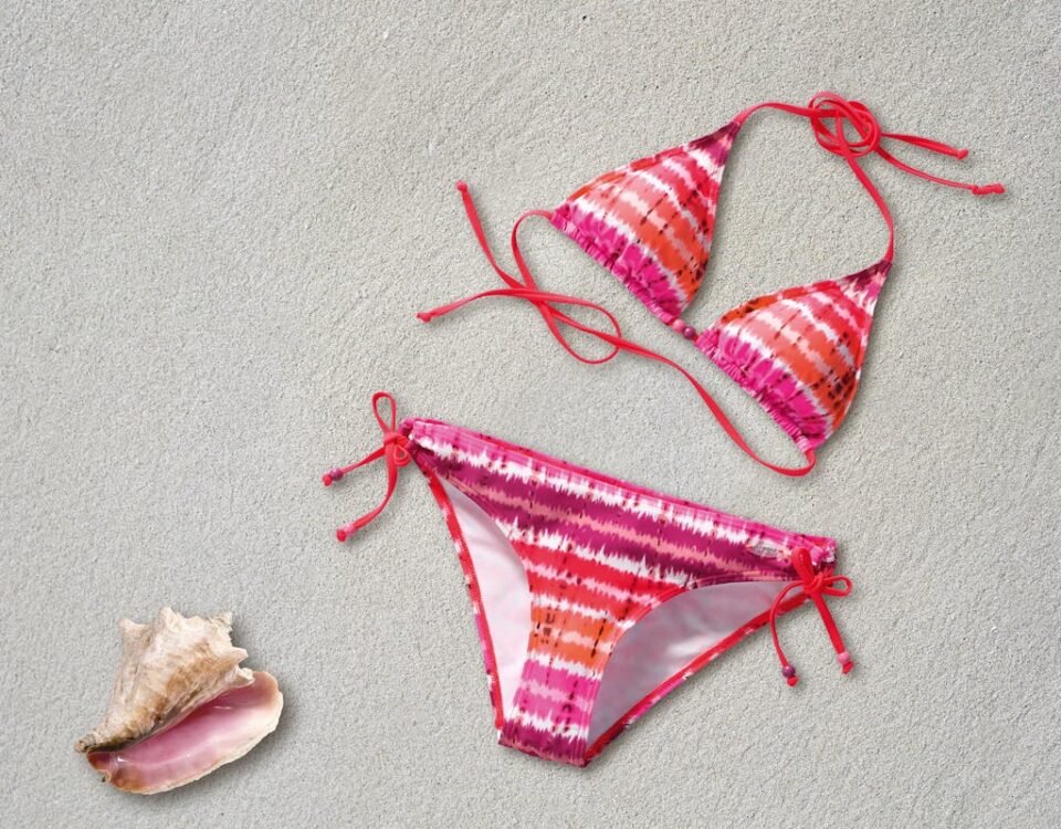Biotecnologia por trás dos tecidos de moda praia Colorful tie-dye bikini on sand next to a pink seashell, ideal for summer vibes.