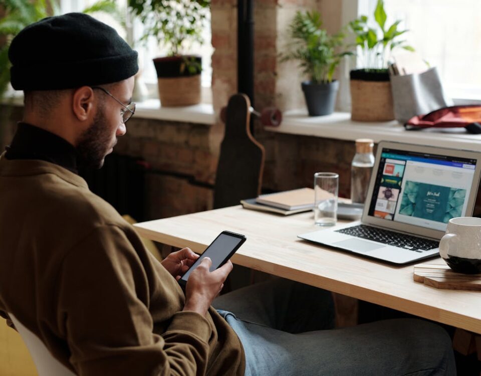 Aplicativos que ajudam no estudo para provas da CNH Male designer working on laptop and phone in a modern home office setting.