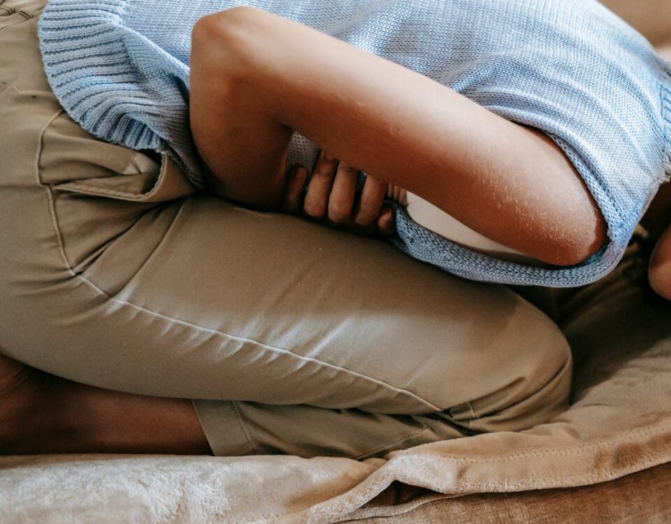 Tecnologias que ajudam no controle de doenças inflamatórias A young woman in discomfort, holding her stomach while curled up on a couch indoors.