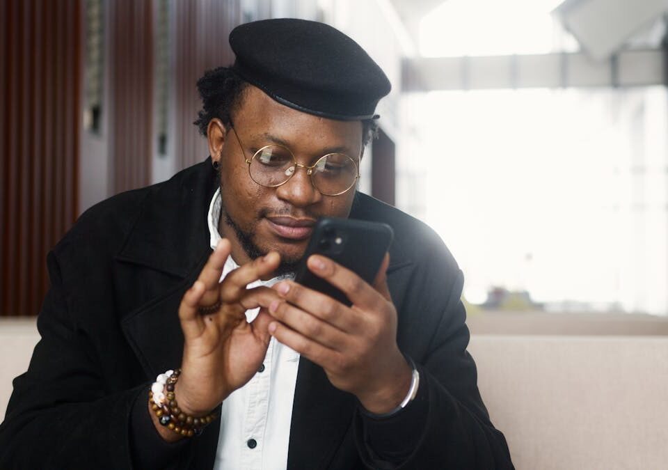 Apps para comparar preços de seguros com facilidade A cheerful man wearing a stylish outfit uses a smartphone while seated in a modern indoor setting.