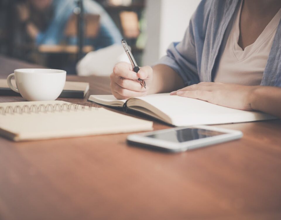 Aplicativos para ajudar a passar no exame teórico da CNH A woman writes in a notebook at a café table with a coffee and smartphone nearby.