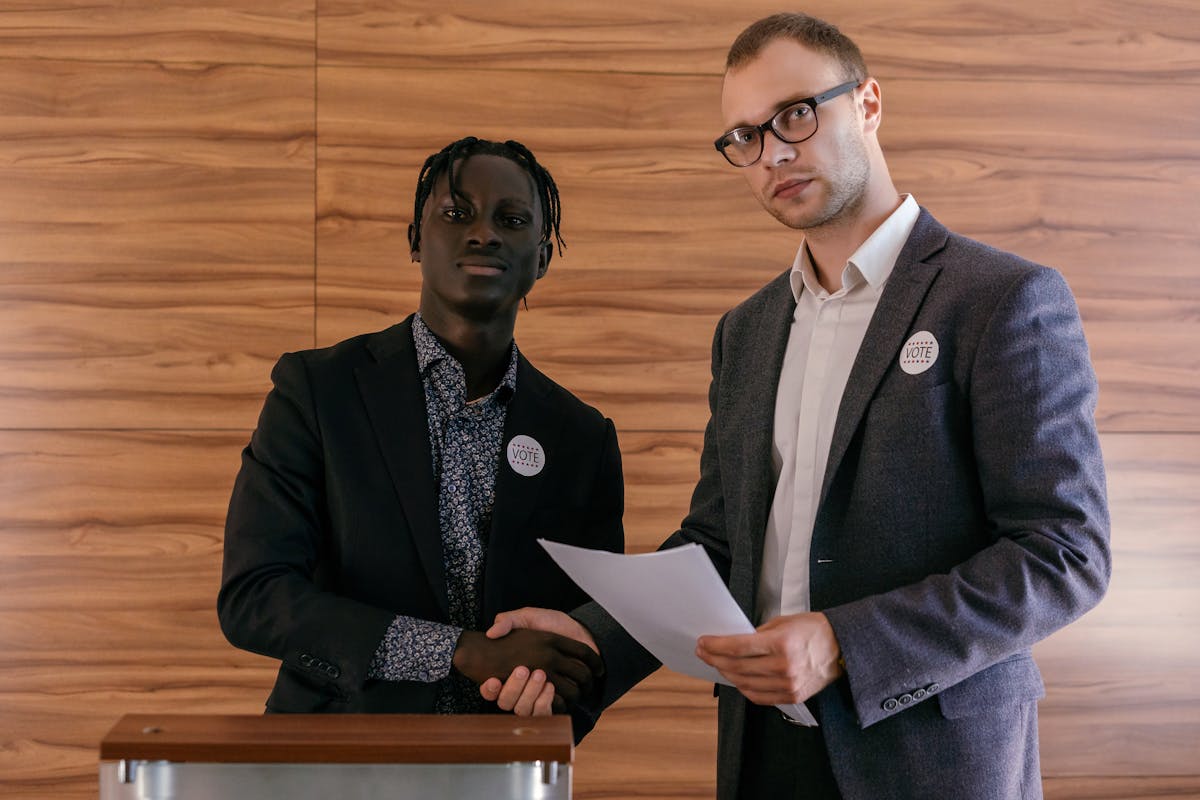 Ferramentas que ajudam a monitorar discursos políticos Two men in business attire shaking hands with agreement, promoting voting indoors.