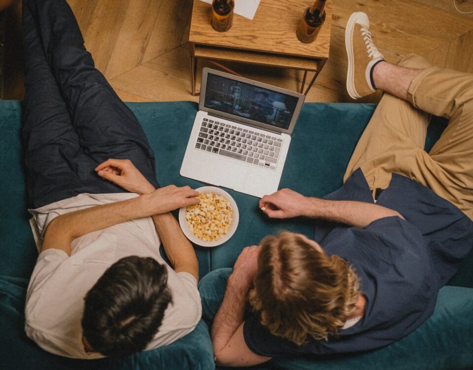 Plataformas de streaming: como escolher a ideal Two friends enjoying a movie on a laptop with popcorn from a cozy couch perspective.