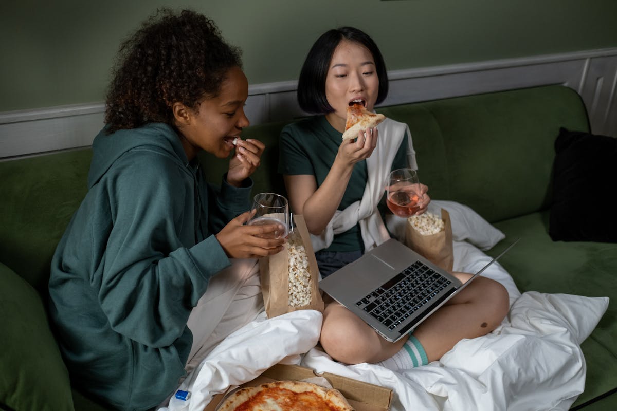 Plataformas de streaming: como escolher a ideal Two women relaxing indoors with pizza, popcorn, and drinks, watching a movie on a laptop.