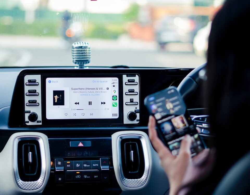 Como funcionam os assistentes virtuais para motoristas Car dashboard with a touchscreen display and person using a smartphone inside a vehicle.