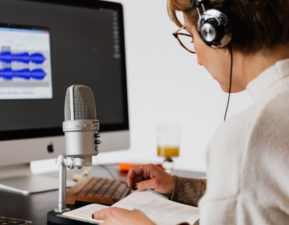 Qual a diferença entre drivers de áudio e codecs? A woman using a microphone and computer to record and edit a podcast while reading from a book.
