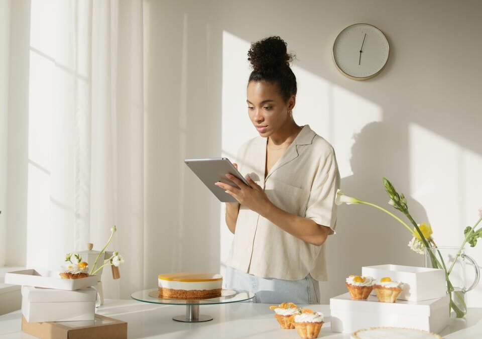 Como a tecnologia melhora suas receitas favoritas A woman using a tablet in a bright and modern kitchen setting, surrounded by cakes and flowers, depicting a lifestyle scene.